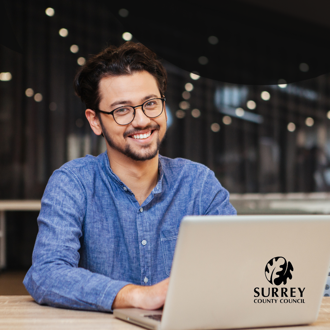 An image of a man, smiling, using a laptop. Surrey County Council logo.